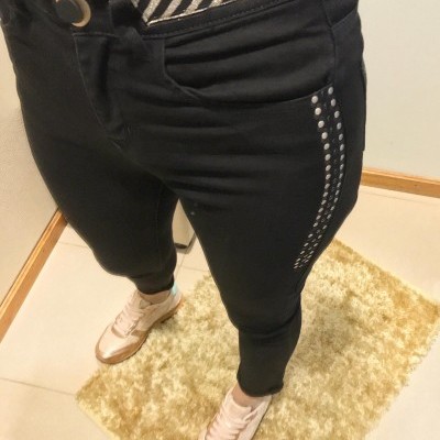 Calças pretas com tachas e cinto riscado prateado, sapatilhas brancas e rosa claro em chão de azulejo com tapete bege