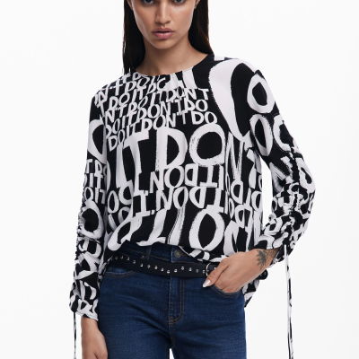 Blusa feminina preta e branca com padrão de texto, mangas longas com laços, jeans azul escuro e cinto preto com tachas.