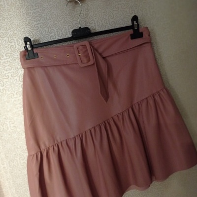 Saia rosa com cinto largo e fivela pendurada num cabide preto contra parede clara com padrão ornamental