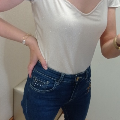 Pessoa vestindo t-shirt branca brilhante de manga curta e jeans azul escuro com detalhes em tachas, sapatos brancos