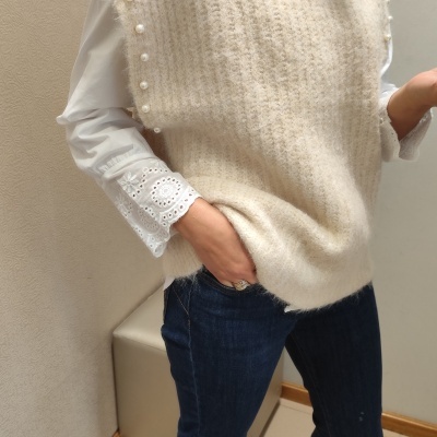 Mulher a usar colete creme de malha com pérolas, camisa branca bordada e calças de ganga azuis.