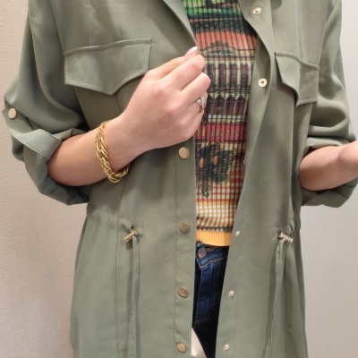 Casaco verde caqui com botões, bolsos no peito e cordão na cintura usado sobre camisola colorida e calças azuis