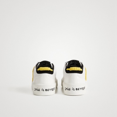 Par de sapatilhas brancas com detalhes amarelos e pretos e texto SMILE IS BETTER na sola
