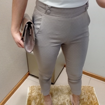 Pessoa com calças cinza de padrão pontilhado, camisa branca, sapatos beges e bolsa bege texturizada sobre tapete bege.