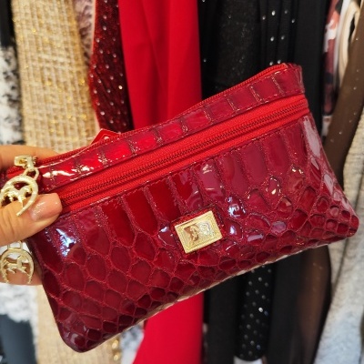 Carteira vermelha brilhante com textura de pele de crocodilo e etiqueta dourada em frente a roupas penduradas