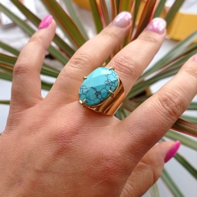 Anel dourado com pedra turquesa em mão com unhas pintadas, fundo com folhas verdes