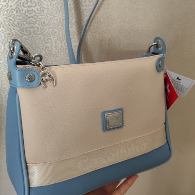 Mala de mão creme e azul claro com logótipo metalizado e alça azul pendurada no gancho