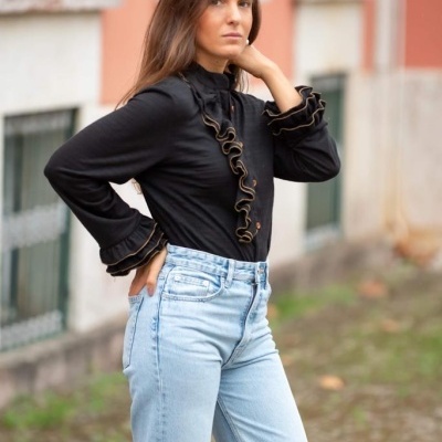 Mulher veste camisa preta com babados dourados e calças de ganga claras ao ar livre