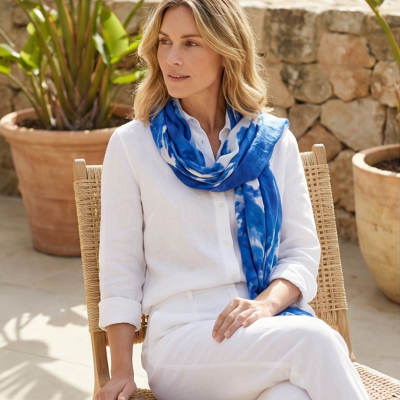 Mulher com lenço azul e roupa branca sentada numa cadeira de madeira com corda em ambiente exterior com vasos e plantas.