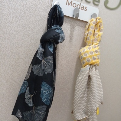 Duas echarpes penduradas num cabide com texto 'Prince Modas', uma preta com folhas cinzentas e outra amarela com padrão circular e tasséis amarelos.