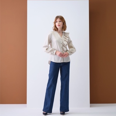 Mulher de pé vestindo camisa bege com folho e calças de ganga azul em estúdio minimalista