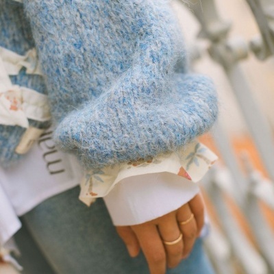 Mão com anéis, camisola azul de malha e camisa branca floral com jeans azul em fundo desfocado