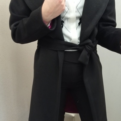 Mulher vestindo casaco preto com cinto sobre camisa branca com padrão preto e calças pretas.