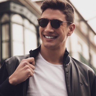 Homem sorridente com óculos de sol, t-shirt branca e casaco preto em rua urbana