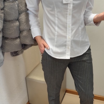 Camisa branca de algodão com botões e calças cinzentas de riscas, botas pretas num interior com tapete bege e casaco peludo cinzento.