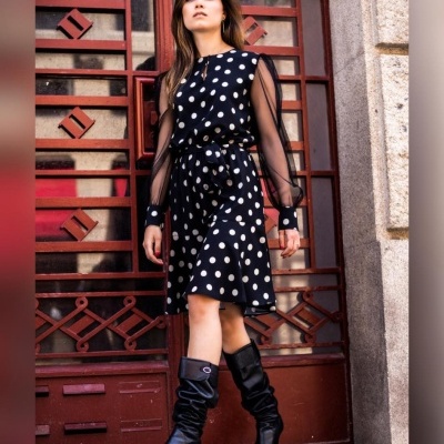 Mulher com vestido preto de bolinhas brancas e botas pretas em frente a porta vermelha