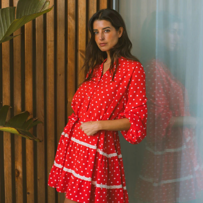 Vestido curto vermelho com bolas brancas e renda, tecido leve, mangas 3/4, fita na cintura, posado junto a parede de madeira e vidro.