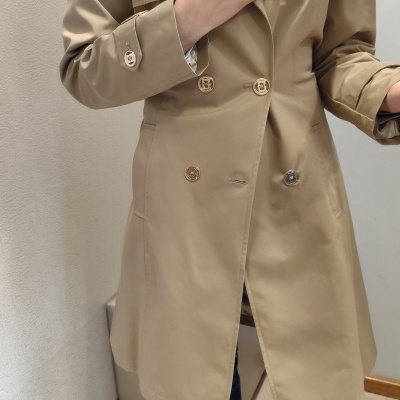 Casaco trench coat bege com botões dourados e prateados, camisa branca, jeans azuis e tapete bege.