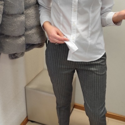 Pessoa vestindo camisa branca com etiqueta SIGNAL woman, calças cinzentas de riscas pretas e botas pretas brilhantes, em ambiente interior com casaco cinzento felpudo ao lado e tapete bege.