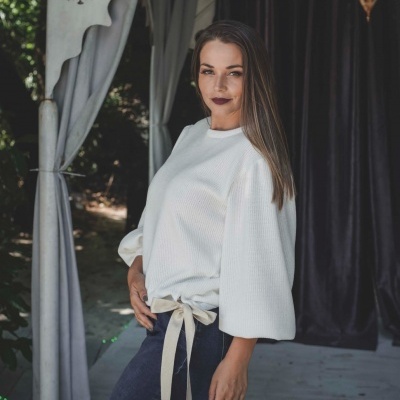 Mulher com túnica branca e calças escuras em ambiente exterior com cortinas