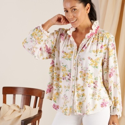 Blusa feminina floral branca com detalhes em renda vestida por mulher, cadeira de madeira e saco de tecido ao fundo.