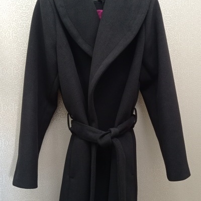Casaco preto comprido com cinto pendurado numa parede clara