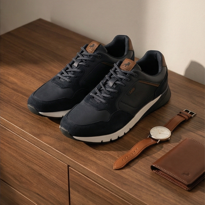 Sapatilhas pretas com detalhes castanhos ao lado de relógio e carteira castanhos sobre móvel de madeira.