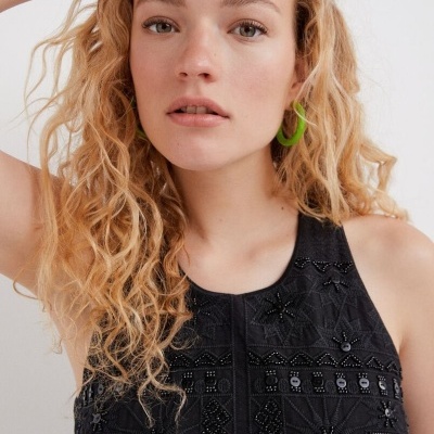 Top preto bordado com lantejoulas e mulher com cabelos loiros encaracolados e brincos verdes