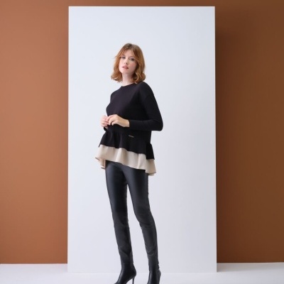 Mulher com casaco preto de malha com barra bege e calças pretas num fundo branco e castanho.