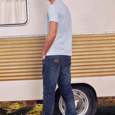 Homem com t-shirt azul clara e jeans azuis escuros junto a trailer branco com faixa amarela