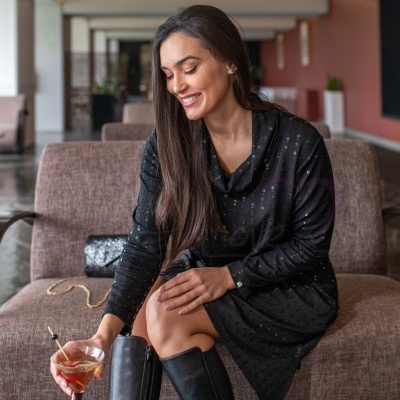 Mulher sorridente sentada num sofá com vestido preto brilhante e botas, segurando cocktail em copo elegante