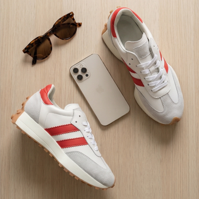 Ténis brancos com riscas vermelhas, óculos de sol castanhos e smartphone branco em superfície de madeira