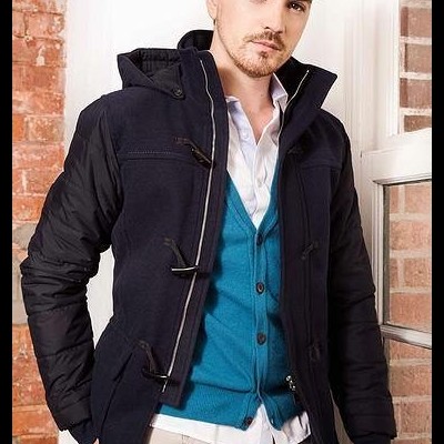 Homem com casaco azul escuro, cardigan azul turquesa, camisa branca e calças bege num ambiente interior com parede de tijolos e janela