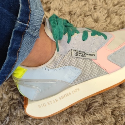 Ténis multicoloridos com sola branca e atacadores verdes, usados com jeans azuis sobre tapete castanho