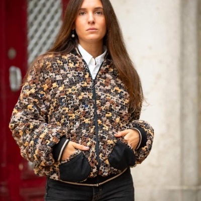 Mulher com casaco texturizado castanho-bege, bolsos pretos, camisa branca e calças pretas, em ambiente urbano.