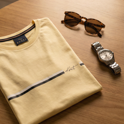 T-shirt amarela com linha decorativa e palavra Koati, óculos de sol e relógio sobre mesa de madeira