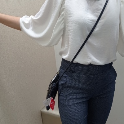 Pessoa com blusa branca de mangas bufantes, calças azul escuras, colar verde e bolsa preta em ambiente interno