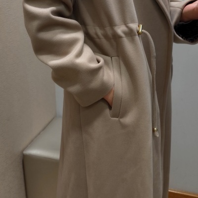 Casaco comprido bege com cinto e gola ampla, usado em ambiente interior