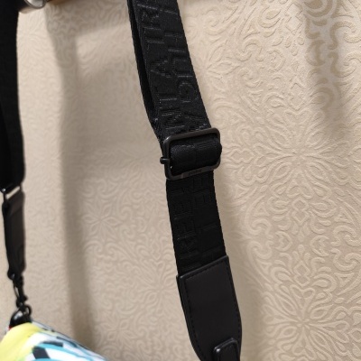 Alça preta ajustável em tecido com letras em relevo numa bolsa colorida e parede bege ao fundo