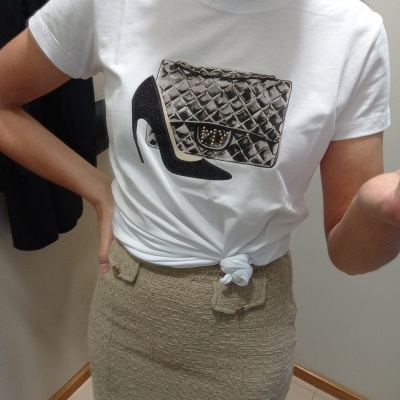 Pessoa a vestir t-shirt branca com estampado prateado de sapato de salto alto e mala acolchoada metálica, saia bege texturada.