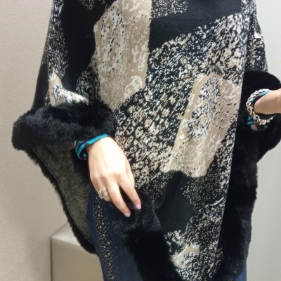 Poncho feminino padrão abstrato com bordas de pele preta, usado com jeans azuis e pulseira prateada.