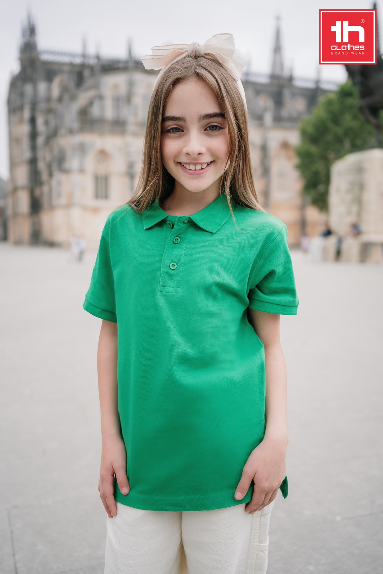 Polo de criança unissexo- ADAM KID Menina de polo verde com laço branco no cabelo em ambiente histórico