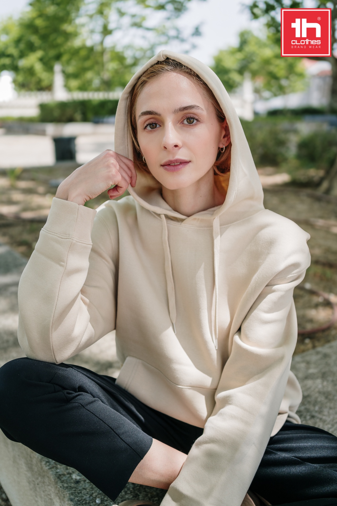 Sweatshirt unissexo com capuz - PHOENIX Mulher jovem com hoodie bege e calças pretas sentada ao ar livre