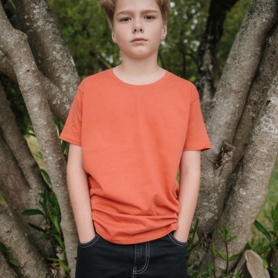 Rapaz com t-shirt laranja e calças pretas junto a uma árvore, logo th Clothes à direita