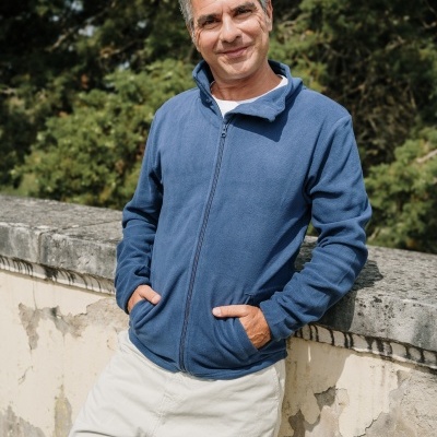 Homem com casaco azul de cinto e calças brancas encostado numa parede de pedra desgastada ao ar livre