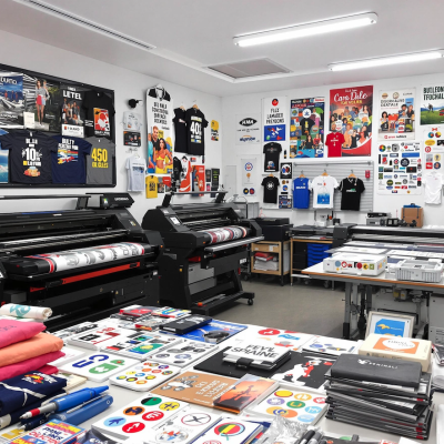 Sala de impressão com máquinas grandes, t-shirts penduradas coloridas e vários produtos gráficos sobre mesas e paredes