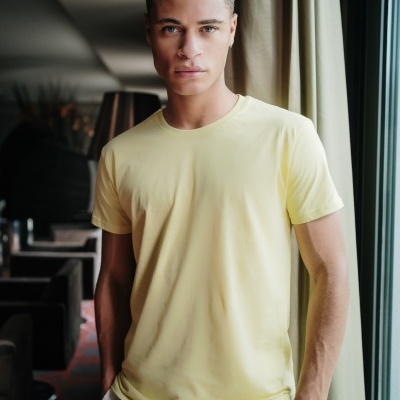 Homem com t-shirt amarela junto a janela com cortinas verdes e logotipo 1h Clothes no topo