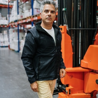 Homem de casaco preto e calças bege em armazém industrial junto a máquina laranja