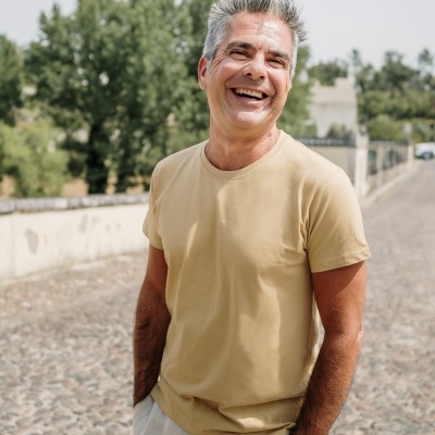 Homem com t-shirt bege e calças claras ao ar livre com árvores e calçada