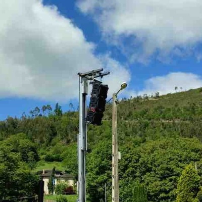 Câmara de vigilância no poste metálico ao lado de poste de iluminação em área verde com colinas e céu azul